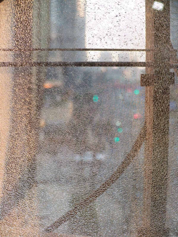 A city street is viewed through a large pane of cracked glass. The cracks create an intricate web-like pattern, partially obscuring the blurred urban lights and buildings visible in the background.
