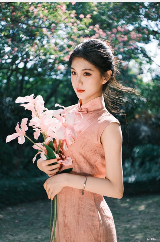 A woman in a sleeveless pink dress stands outdoors holding a bouquet of pink flowers, with sunlight filtering through trees in the background. She looks slightly to the side with a calm expression.