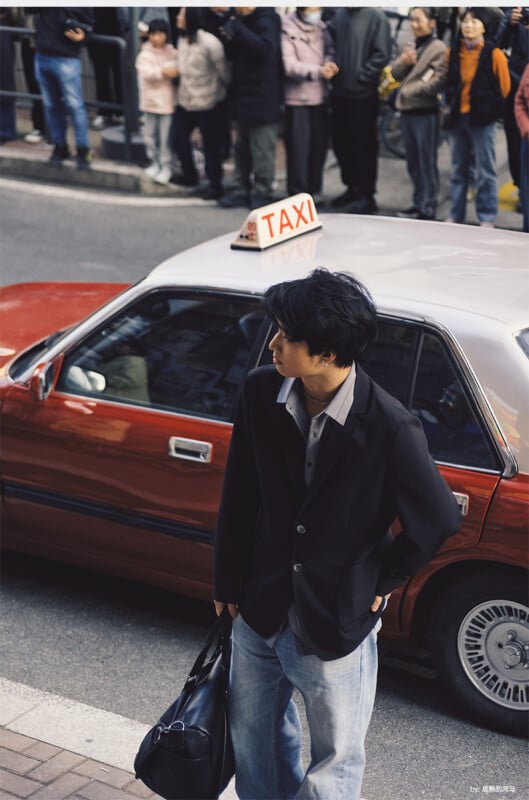 A man in a black blazer and jeans stands next to a red taxi on a city street, holding a black bag. A crowd of people waits on the sidewalk in the background.