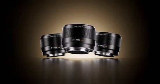 Three camera lenses labeled AF 24mm, AF 50mm, and AF 85mm are displayed on a dark reflective surface with a warm gradient background, positioned upright with the 50mm lens in the center.