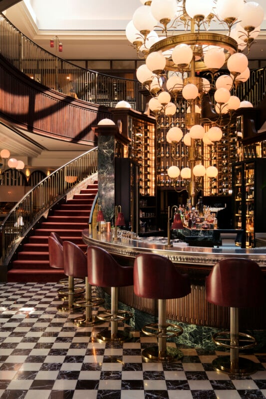 El lujoso bar cuenta con un mostrador curvo, taburetes de cuero rojo, una gran lámpara de araña con luces tipo globo, detalles en mármol negro, un piso de tablero de ajedrez y una gran escalera con una alfombra roja y una barandilla dorada al fondo.