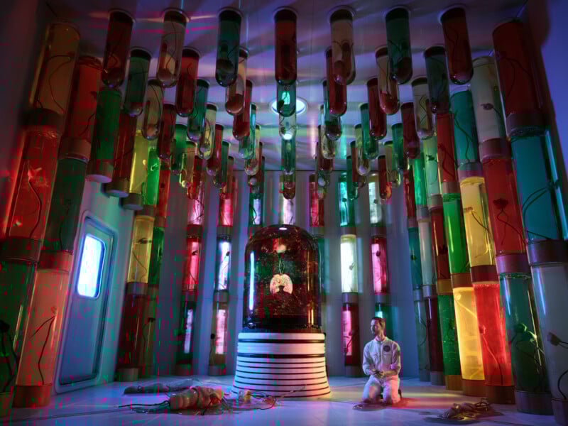 Un hombre vestido de blanco se arrodilla en el suelo de una habitación colorida y futurista llena de tubos brillantes que contienen objetos extraños. En el centro se encuentra una gran cúpula de cristal que contiene un ejemplar rodeado de luces vibrantes.