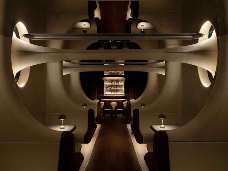 El interior del bar es futurista y tiene poca iluminación, con áreas curvas para sentarse, pequeñas mesas redondas con luces y una barra central brillante en la parte trasera, rodeada de paredes esculturales y una cálida iluminación ambiental.