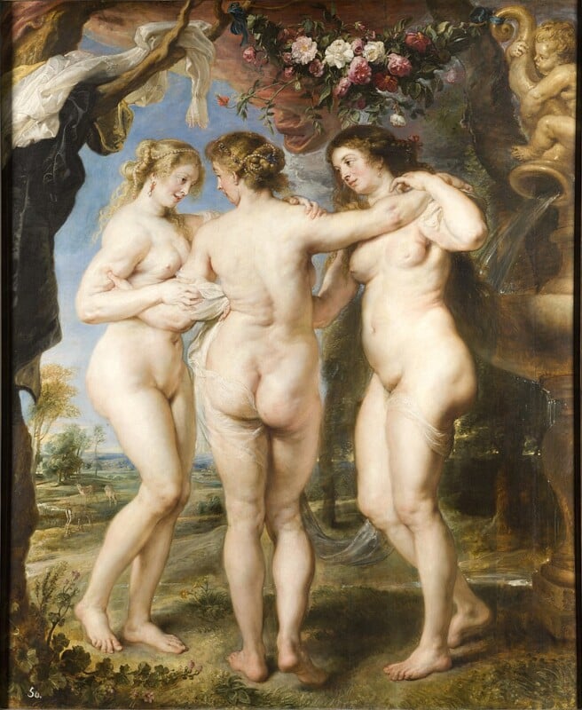 Three nude women stand close together outdoors under a garland of flowers, embracing each other. The scene has a classical, mythological feel, with a cherub statue and lush landscape in the background.