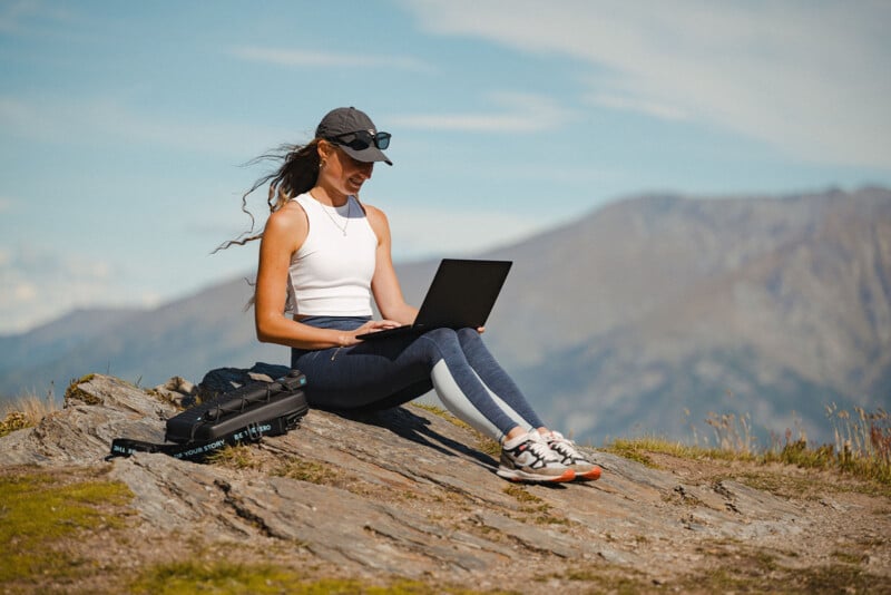 Una mujer con ropa deportiva se sienta en una pendiente rocosa al aire libre y usa una computadora portátil. Lleva sombrero, gafas de sol y zapatillas de deporte con montañas y cielo azul de fondo. Una bolsa negra yacía sobre la roca junto a ella.