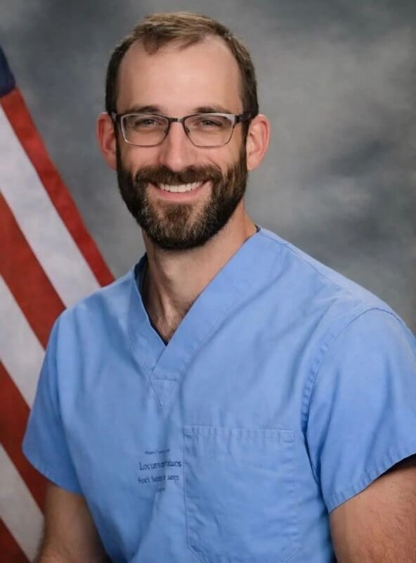 A man with glasses and a beard, smiling, wearing light blue medical scrubs, sits in front of a gray background with part of an American flag visible on the left.