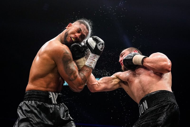 Two male boxers in a ring, both wearing black trunks, as one lands a powerful punch to the other's face, causing sweat to fly and the recipient to grimace and recoil from the impact.