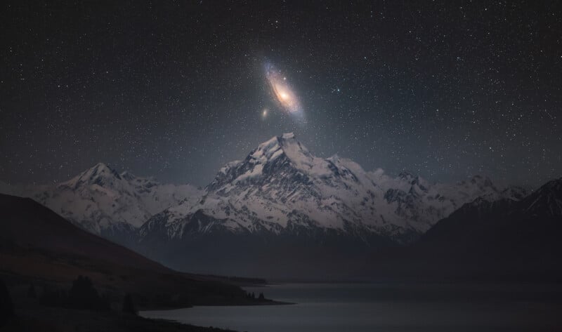 Snow-capped mountains under a starry night sky, with a bright spiral galaxy and glowing core appearing just above the tallest peak, reflected in a calm lake below.