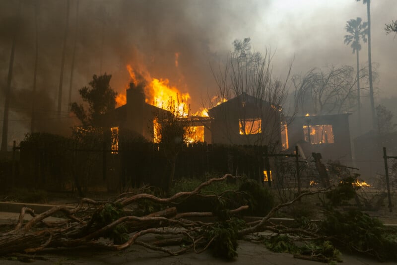 A house engulfed in large flames and thick smoke during a wildfire, with fallen branches and debris scattered on the ground in front. Trees and vegetation around the house are also burning.