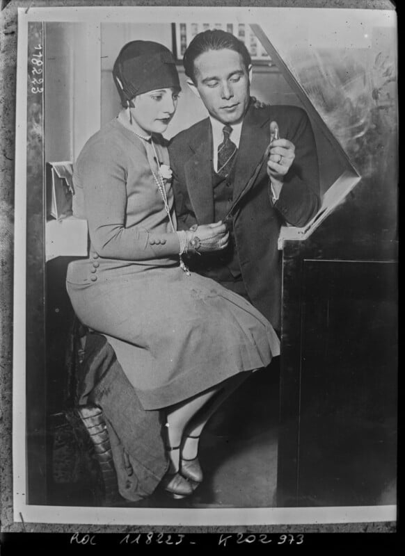 A woman in a cloche hat and tailored dress sits beside a man in a suit. They both look at a small object he is holding, possibly a key or pen. The photo appears vintage, likely from the early 20th century.