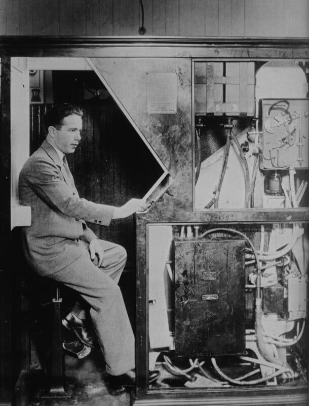 A man in a suit sits inside an old-fashioned photo booth, holding up a piece of paper. The booth’s mechanical and electrical components are exposed and visible beside him. The photo is black and white.