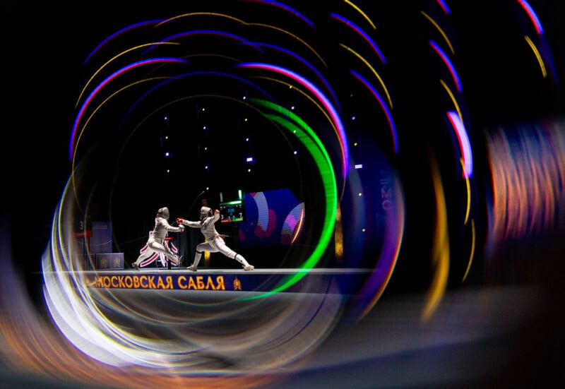 Two fencers in white uniforms duel on a lit stage, surrounded by colorful, swirling light effects; Russian text is visible on the platform beneath them.