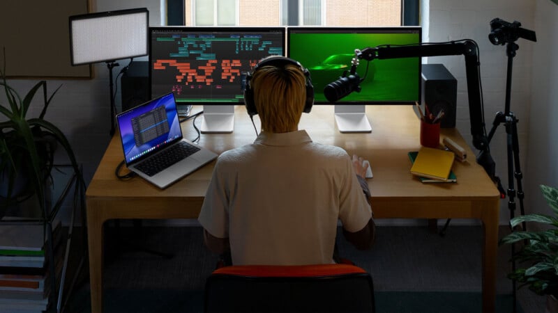 A person with headphones sits at a desk using a laptop and two monitors. The desk also has a microphone, camera on a tripod, speakers, notebooks, and plants. The screens show code and an underwater scene.