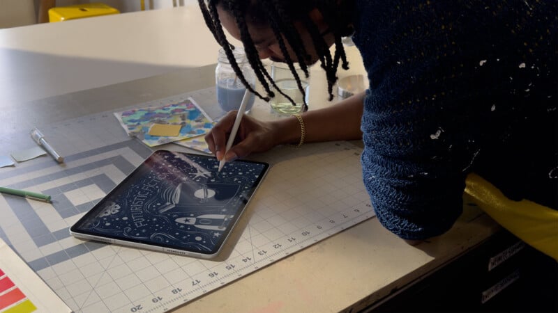 A person with braided hair uses a stylus to draw a futuristic robot face on a tablet, leaning over a gridded worktable with art supplies and colorful paper nearby.