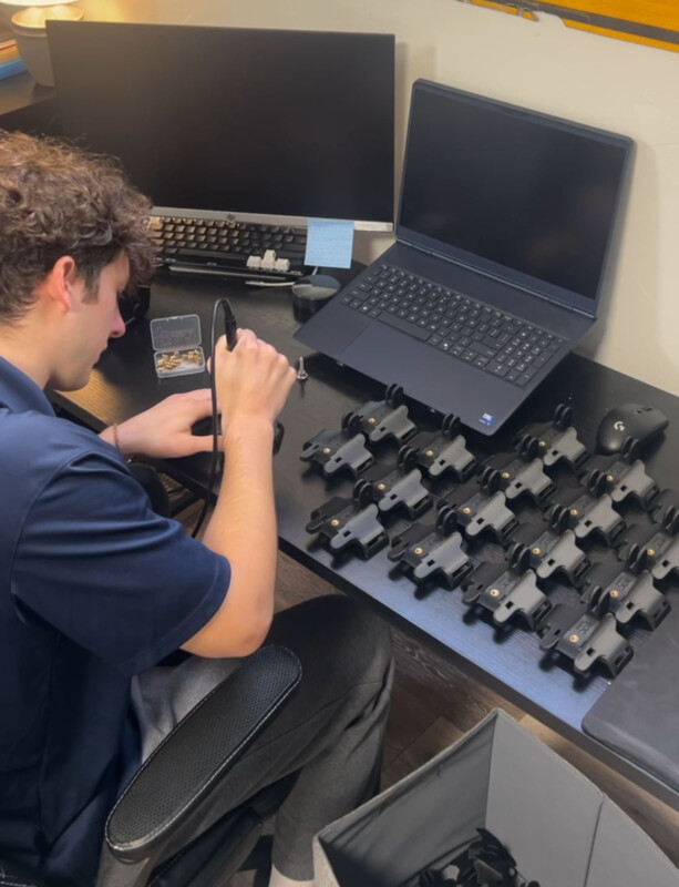 Un hombre está sentado en un escritorio y usa herramientas para ensamblar múltiples dispositivos electrónicos negros. Sobre la mesa hay una computadora portátil, un monitor de computadora y cajas de piezas pequeñas, así como una fila de dispositivos idénticos en uso.