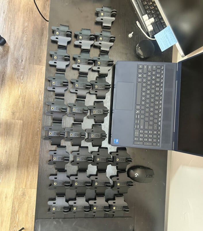 Veinticuatro estuches idénticos de cuero y plástico negro están alineados en fila sobre una mesa negra, junto a computadoras portátiles, ratones y monitores. Se ven claramente la mesa y parte del suelo de madera.