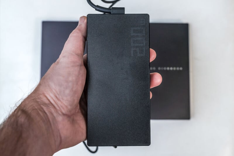 A hand holding a large, black rectangular power adapter with a textured surface and "200" printed on it; a closed black laptop is visible in the background.