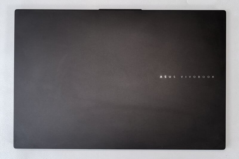 A closed, black ASUS VivoBook laptop is shown from above on a light, textured surface, with the logo positioned off-center to the right.