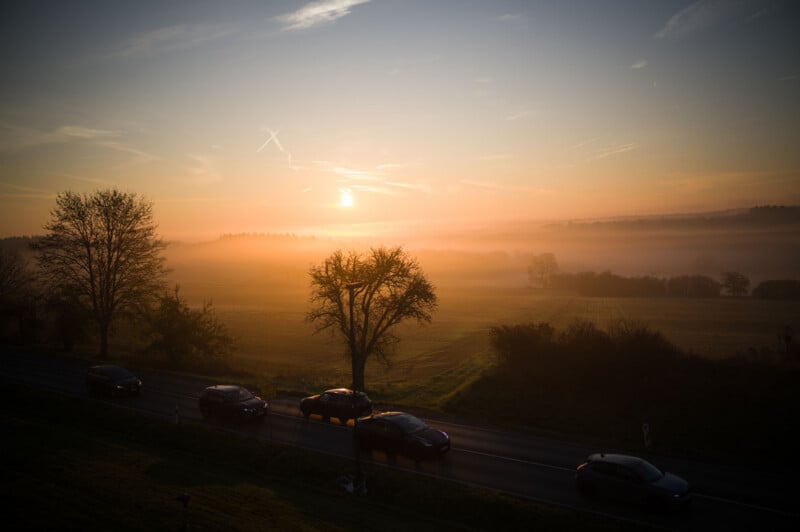 Cars drive along a road bordered by trees at sunrise, with mist covering the fields in the background and a golden sky illuminating the landscape.