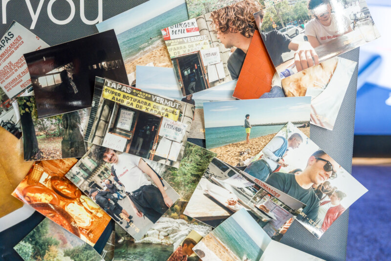 A scattered pile of colorful printed photographs, mostly showing people smiling outdoors, food, beach scenes, and a shop with a “TAPAS Y VERMUT” sign. The photos overlap on a dark surface.