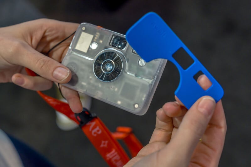 A person holds a transparent camera in one hand and a blue plastic camera cover in the other. A red lanyard is attached to the camera. The camera’s inner components are visible through its clear case.