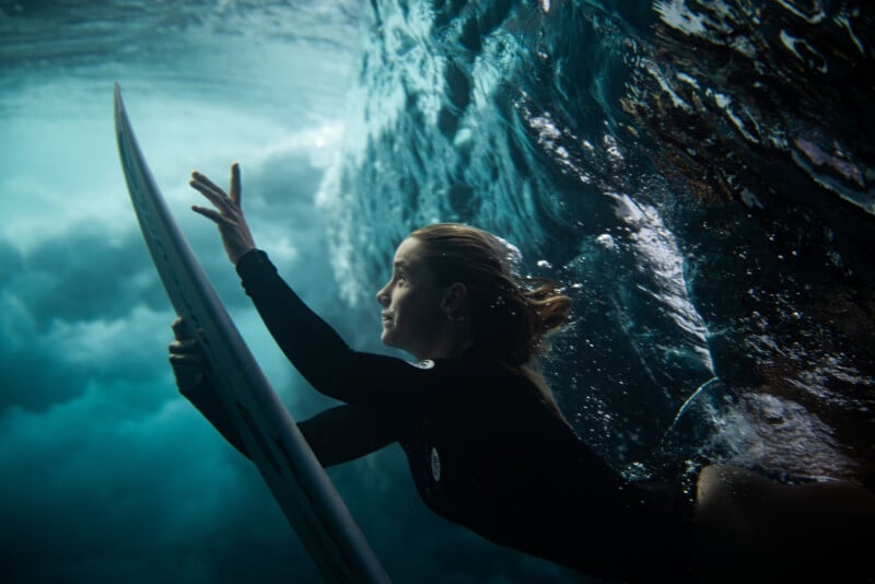 A person in a black wetsuit swims underwater while holding a surfboard, surrounded by swirling blue water and bubbles, with light filtering through the waves above.