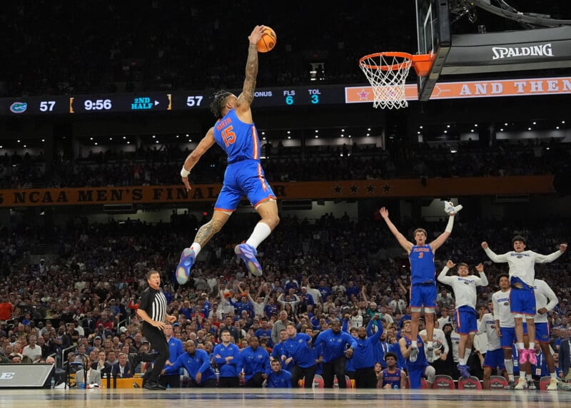 A basketball player in a blue uniform leaps toward the hoop for a dunk as teammates and fans cheer. The scoreboard shows a tied game, and the opposing team watches from the court. The arena is packed with spectators.