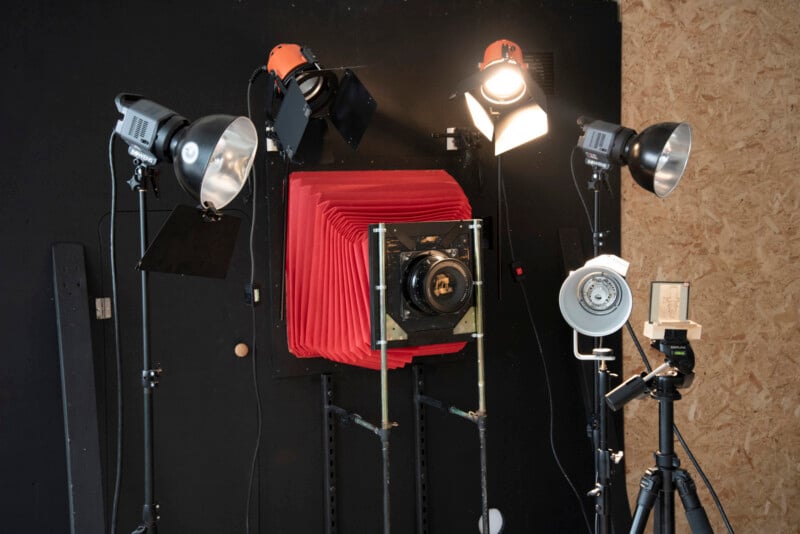 A large-format camera with a red bellows is set up in a studio, surrounded by multiple professional studio lights, reflectors, and a tripod on a wooden floor with a black and wood-paneled background.