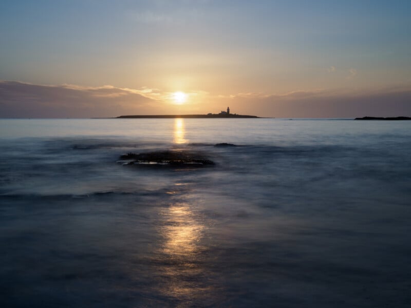 A calm seascape at sunset, with the sun low on the horizon casting a golden reflection across the water. A distant island with a small lighthouse is silhouetted against the sky.
