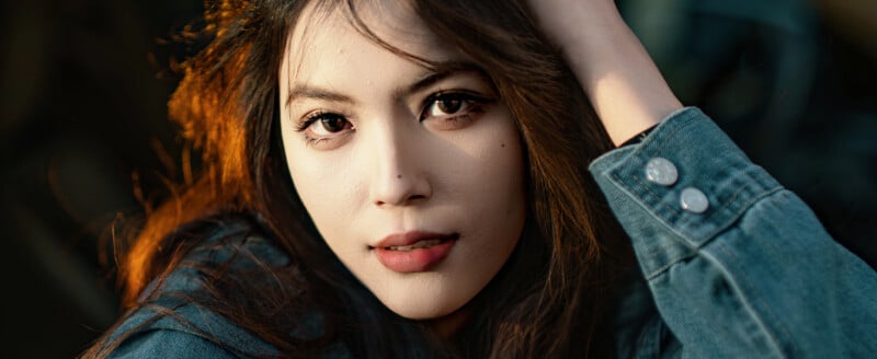 Primer plano de una mujer joven con cabello largo y castaño, vestida con una chaqueta vaquera, mirando directamente a la cámara con una expresión relajada y tocándose la cabeza con una mano, bajo una cálida luz natural.