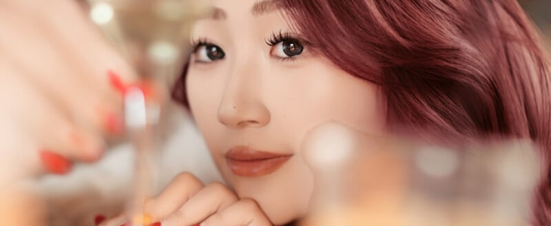 Primer plano de una mujer con cabello rojo largo y ondulado y maquillaje natural sosteniendo un vaso y mirando a la cámara. Una iluminación suave y cálida crea un ambiente confortable y festivo.