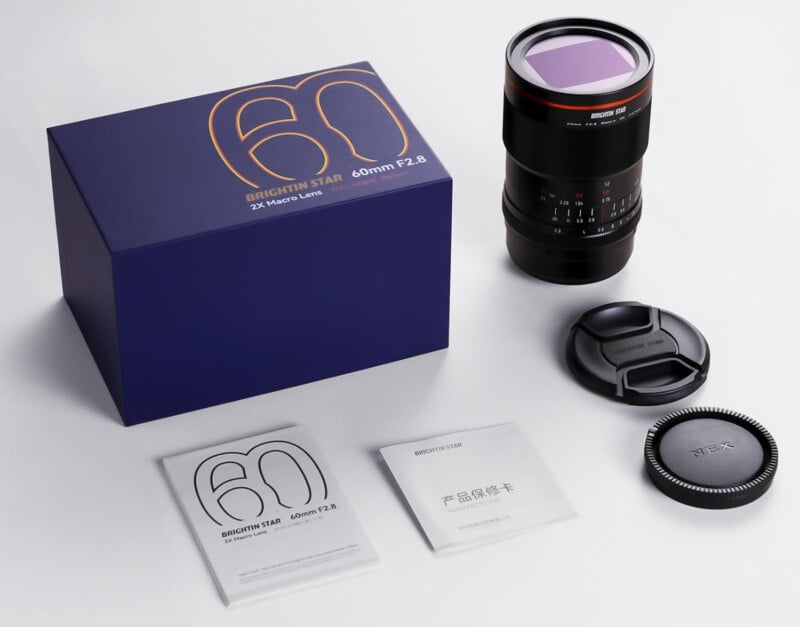 A 60mm f/2.8 camera lens with lens caps, a blue product box, and two instruction booklets are displayed on a white surface. The lens has red accents and the box features an orange "60" design.