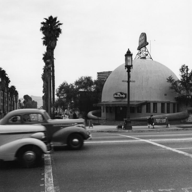A black and white photo of a street with classic 1940s cars driving past the round, domed building of The Dome Drive-In Dairy. Palm trees and a tall street lamp line the road; a large sign sits atop the dome.