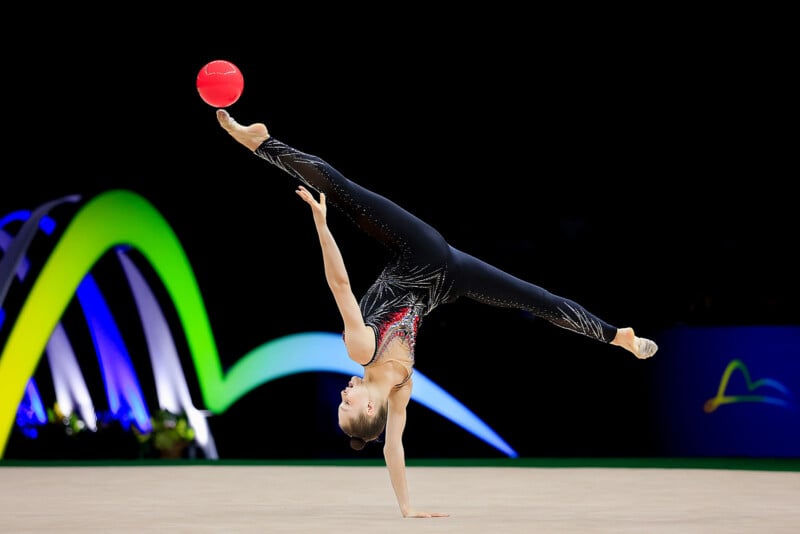 A rhythmic gymnast performs a one-handed handstand while balancing a red ball on her raised foot, with colorful curved lights in the background.