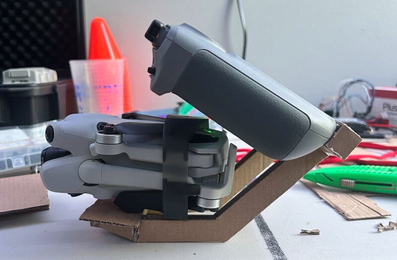 Un dron plegado y su controlador se colocan sobre un soporte de cartón personalizado, rodeados de herramientas y materiales de bricolaje, incluido un cuchillo verde y un cono de plástico naranja, en un espacio de trabajo desordenado.