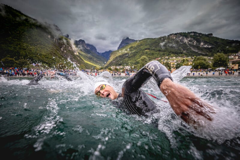 A swimmer in a wetsuit and goggles competes in an open water race, arm outstretched mid-stroke, with mountains and a cloudy sky in the background. Crowds line the shore.
