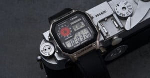 A Casio digital wristwatch with a black strap and multiple displays rests on top of a vintage silver camera on a dark surface. The watch face shows the time as 10:03:20 and features world time and illuminator functions.