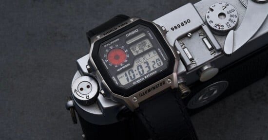 A Casio digital wristwatch with a black strap and multiple displays rests on top of a vintage silver camera on a dark surface. The watch face shows the time as 10:03:20 and features world time and illuminator functions.