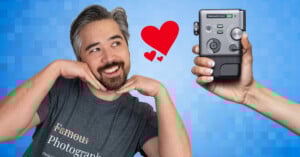 A smiling person in a "Famous Photographer" shirt poses with hands under their chin, looking at a hand holding a camera device. A red heart floats between them on a blue pixelated background.