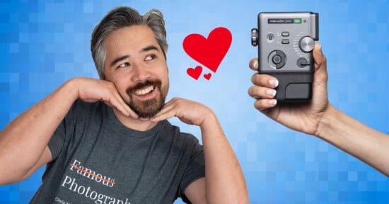 A smiling person in a "Famous Photographer" shirt poses with hands under their chin, looking at a hand holding a camera device. A red heart floats between them on a blue pixelated background.
