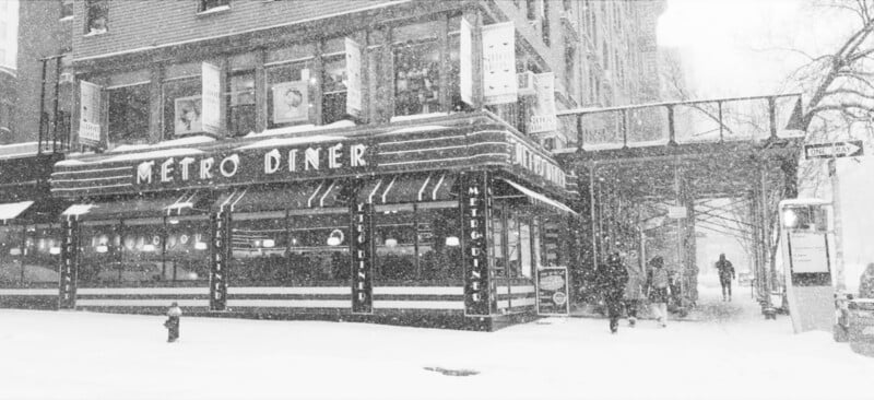 Black-and-white photo of Metro Diner on a snowy city street. Snow covers the ground and buildings, with a few people walking nearby. An elevated walkway connects buildings above the street.