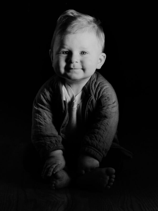 A young child with short light hair sits barefoot on the floor, smiling softly. The image is in black and white with dramatic lighting, casting shadows and highlighting the child’s face and hands.