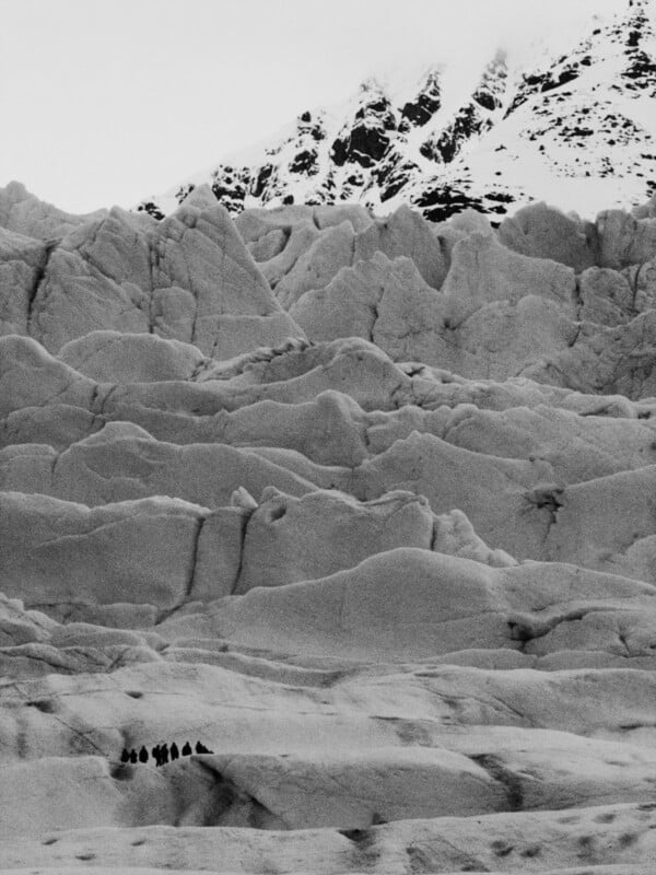 A group of people is dwarfed by a vast, rugged, snow-covered glacier with jagged peaks rising in the background, depicted in a dramatic black and white photograph.