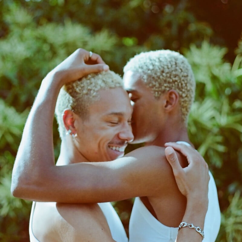 Two people with short, bleached blonde hair embrace closely outdoors, smiling and appearing relaxed and affectionate. Green foliage is softly blurred in the background. Both wear white tops.
