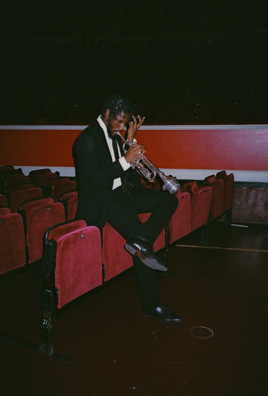 A man in a black suit sits on the armrest of a red theater seat, holding a silver trumpet and adjusting his hand, with empty theater seats around him.