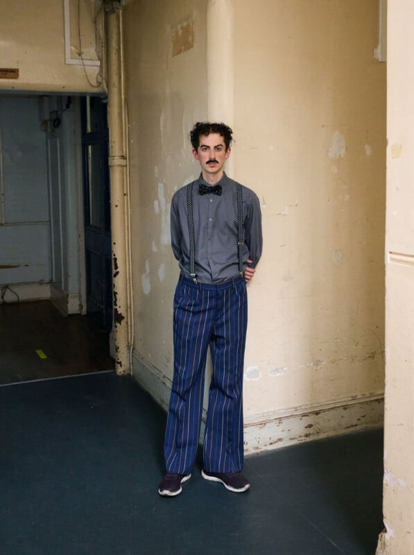 A man with curly hair and a mustache stands indoors against a worn beige wall. He wears a grey shirt, suspenders, a bow tie, and blue pinstriped pants, with his hands behind his back.