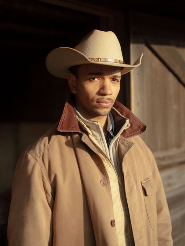 A man wearing a tan cowboy hat and a brown coat stands in front of a wooden structure, looking directly at the camera with a serious expression. Warm sunlight highlights his face and clothing.