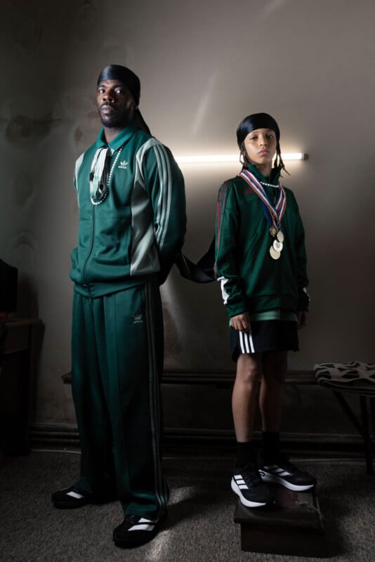 A man and a child wearing green tracksuits and black durags stand indoors. The man has medals and chains around his neck, while the child stands on a box, also wearing medals. A fluorescent light glows behind them.