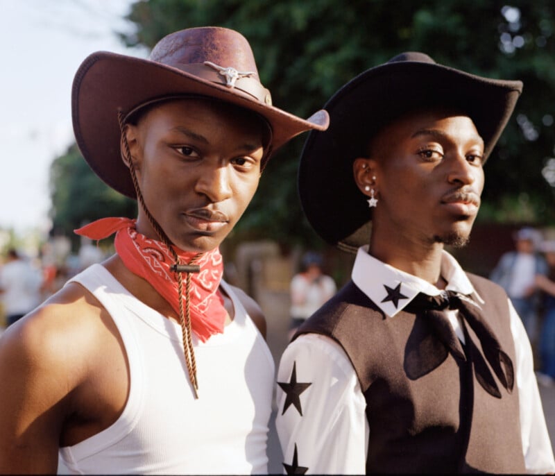 Two young Black men dressed in cowboy hats and western-style outfits, one wearing a red bandana and white tank top, the other in a black vest with white stars and a necktie, standing outdoors with trees in the background.