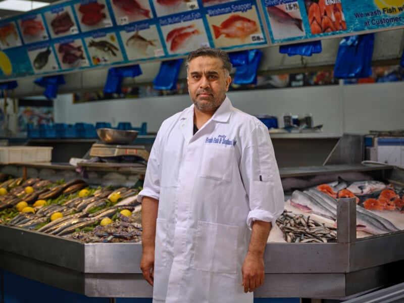 A man in a white coat stands in front of a seafood counter displaying various fresh fish and seafood on ice and lemons, with fish signs and prices above him in a market.